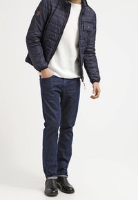 Uomo che indossa una giacca trapuntata blu navy, un maglione bianco, jeans scuri e stivali in pelle neri, in piedi con una mano in tasca.