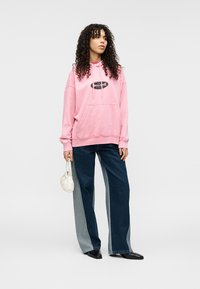 Ροζ φούτερ με κουκούλα, μπροστινή τσέπη καγκουρό και λογότυπο, συνδυασμένο με τζιν παντελόνια φαρδιάς γραμμής με σχέδιο με μπλοκ χρωμάτων.