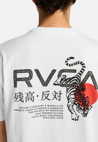 Camiseta de algodón blanca con un gráfico negro de un tigre, un círculo rojo y texto. El diseño incluye caracteres chinos y frases en varios idiomas.