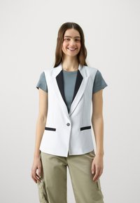 Biely blazer s čiernym klopom a vreckami, zapínanie na jeden gombík, nosený cez svetlomodrú košeľu, v kombinácii s olivovozelenými cargo nohavicami.