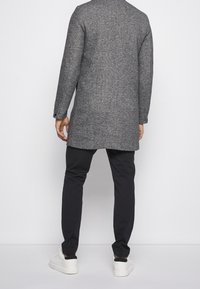 Grau melierter Wollmantel mit langen Ärmeln und Rückenschlitz, kombiniert mit schwarzen Nadelstreifenhosen und weißen Turnschuhen.