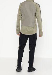 Olivgrüne Sportjacke mit kontrastierendem grauem Rückenteil, langen Ärmeln und schwarzen Hosen mit seitlichen Streifen und strukturierten Akzenten.