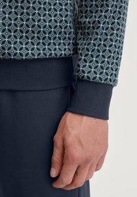 Langärmliges gemustertes Sweatshirt in Dunkelblau und Türkis, mit geometrischen Designs und gerippten Bündchen, kombiniert mit dunklen Hosen.