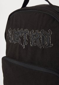 Svart kordflosseryggsäck med en broderad design i silvertråd, med dragkedjestängning på framsidan. Texturerad yta.