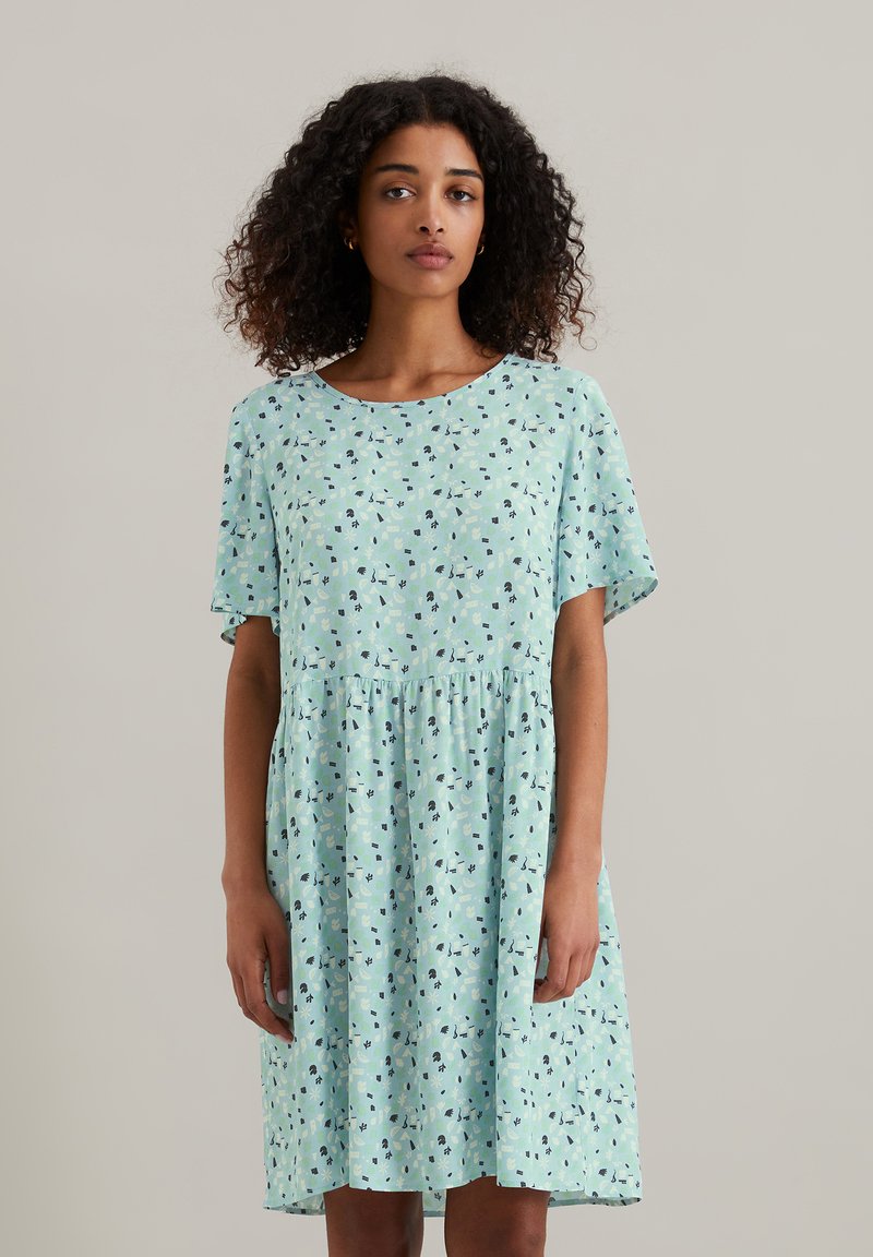 Lichtblauwe jurk met korte mouwen, ronde halslijn, gecraagde taille en een bloemen/geometrisch patroon in donkere kleuren erdoorheen. Zachte stof.