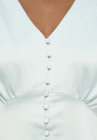 Κοντινό πλάνο ενός γαλάζιου ενδύματος με V-λαιμόκοψη και μια κάθετη σειρά από επτά κουμπιά καλυμμένα στο κέντρο του μπροστινού μέρους.