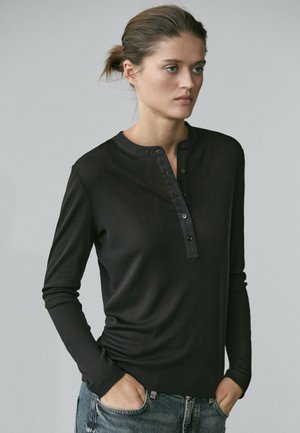 Woman with light skin and tied-back hair wearing a black long-sleeve henley shirt and blue jeans, standing with hands in pockets.