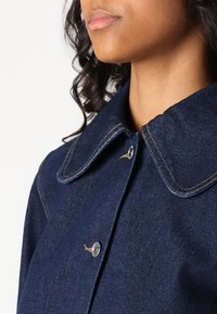 Veste en denim bleu foncé avec un large col, des boutons en argent et des détails de couture visibles. Le tissu a une texture lisse.