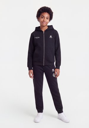 Jeune personne debout portant un sweat à capuche zippé noir et un pantalon de survêtement noir avec des baskets blanches sur fond blanc.