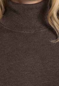 Gros plan d'une personne portant un pull à col roulé marron côtelé avec des mèches de cheveux blonds visibles sur le côté.