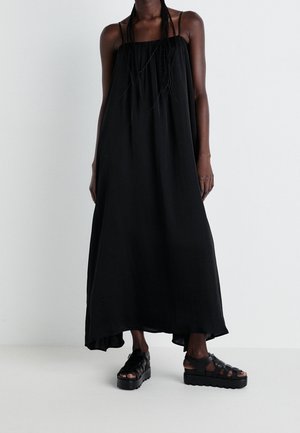 Persona alta con un vestido maxi negro holgado y sin mangas con tirantes finos y sandalias negras de plataforma sobre un fondo blanco.