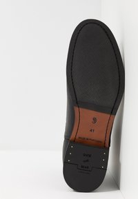 Musta nahkakengän pohja, jossa on teksturoitu musta kumipinta ja ruskea nahkaketju. Sisältää "Shoe the Bear" -brändäyksen ja kultaisia niittejä. Koko 41.