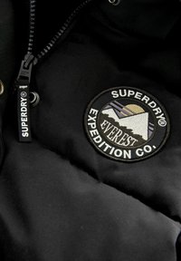 Črna podložena jakna z okroglo vezeno nalepko z grafiko gora in sonca ter blagovno znamko "SUPERDRY" na zadrgi.