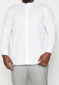 Vit långärmad skjorta med knäppning och mandarin-krage, med en slät, vävd struktur och rak nederkant, tillsammans med grå rutiga byxor.