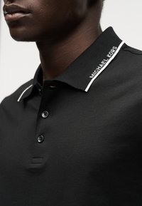 Gros plan d'une personne portant un polo noir avec « Michael Kors » imprimé en blanc sur le col et un passepoil blanc sur l'épaule.