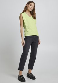 Top senza maniche verde chiaro con scollatura a V, vestibilità rilassata, tessuto testurizzato; abbinato a jeans neri a vita alta e mocassini neri con dettagli in oro.