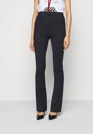 Femme portant un pantalon noir ajusté taille haute avec des coutures à l'avant et des talons noirs pointus, debout devant un fond clair uni.