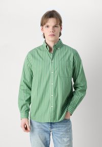 Πράσινο ριγέ πουκάμισο με κουμπιά από ελαφρύ ύφασμα, διαθέτει θήκη στο στήθος και κλασικό γιακά, συνδυασμένο με απαλό τζιν σε ανοιχτό χρώμα και φθορές.