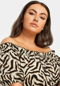 Blusa off-the-shoulder in beige chiaro con una stampa zebrina nera e marrone. Presenta un colletto arricciato e bottoni marroni lungo la parte anteriore.