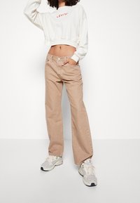 Pantalones vaqueros de pierna ancha beige, hechos de mezclilla, con bolsillos frontales y una cintura de altura media. Combinados con un sudadera corta blanca y zapatillas.