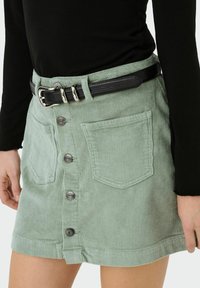 Mini-jupe en velours côtelé vert clair avec boutons devant, poches appliquées et ceinture noire texturée portée par une personne vêtue d'un haut noir à manches longues.