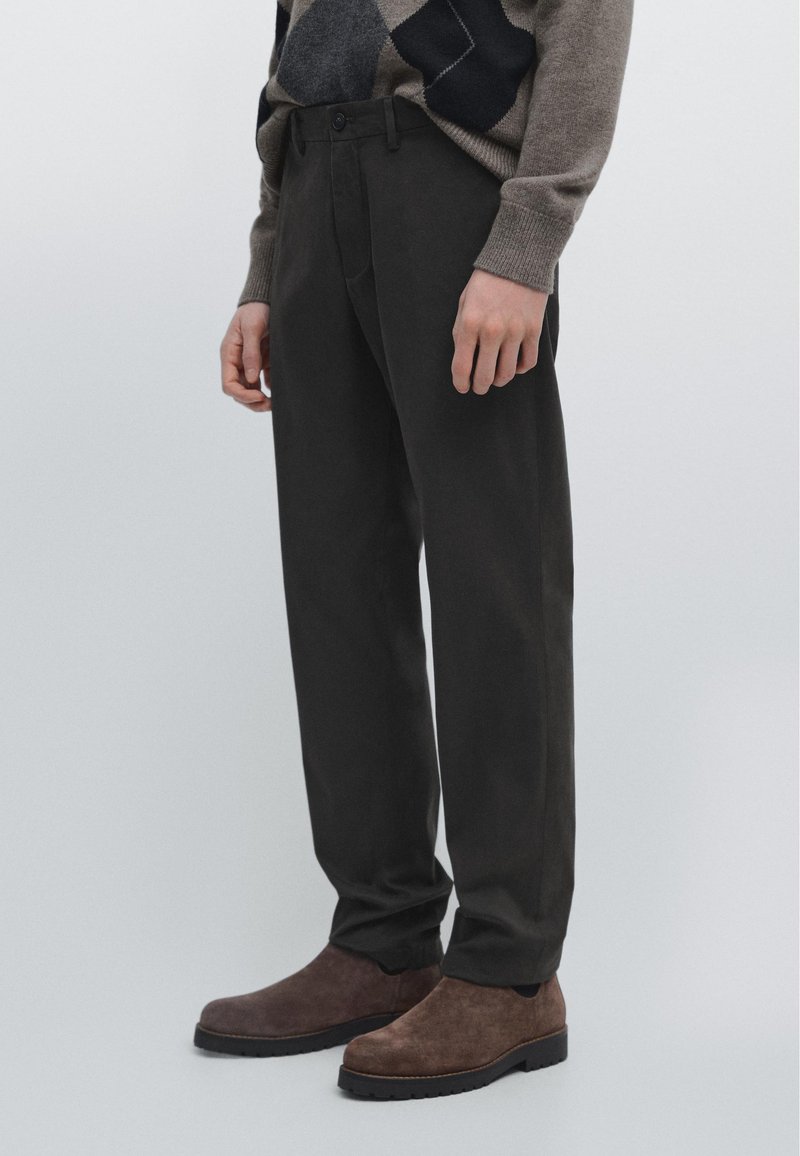 Mannequin portant un pantalon droit marron foncé, des bottes en daim marron, et un pull beige et bleu marine à motif losanges sur un fond clair uni.