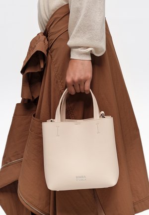 Mano sosteniendo un pequeño bolso beige con el texto "BIMBA Y LOLA", vistiendo una blusa de manga larga beige y una falda marrón con detalle de cremallera.