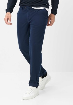 Mann, der dunkelblaue Hosen, weiße Turnschuhe und einen marineblauen Pullover trägt, steht mit einem Bein vor das andere gekreuzt auf weißem Hintergrund.