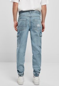 Lyseblå cargo bukser med elastiske manchetter, der har to baglommer og et logopatch. Materialet ser ud til at være denim med en afslappet pasform.