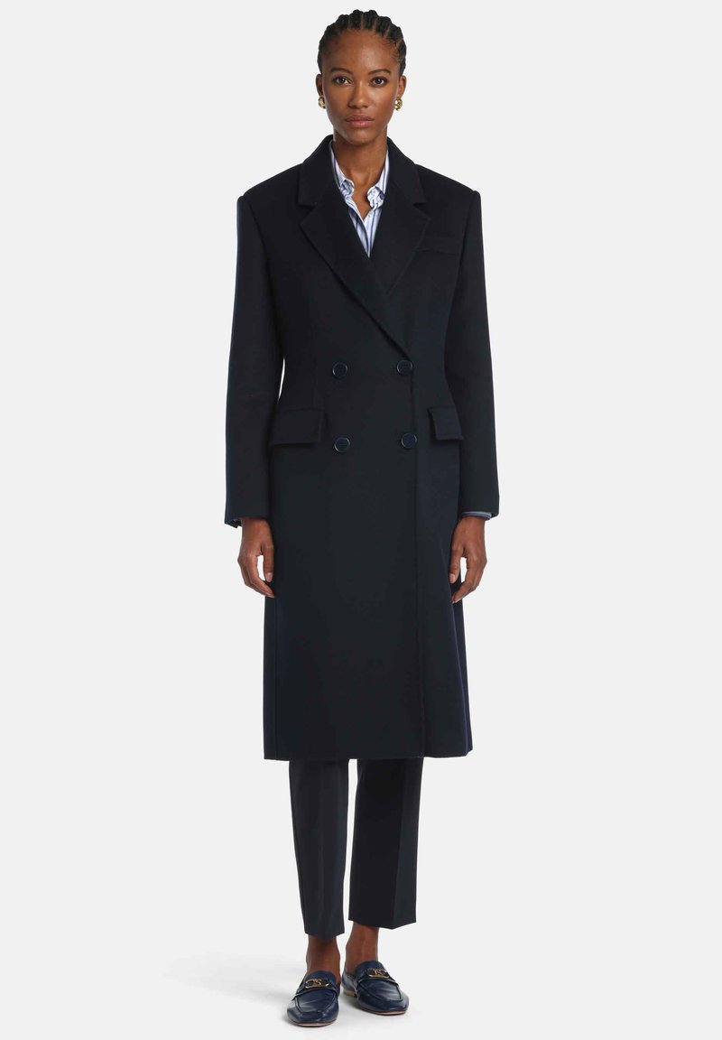 Manteau croisé bleu marine avec larges revers, poches latérales et tissu texturé, associé à un pantalon ajusté et des mocassins.