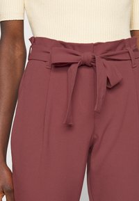 Burgundské kalhoty s vysokým pasem a zavázaným opaskem, vyrobené z hladké látky. Obsahují sklady a boční kapsy pro zvýšení funkčnosti.