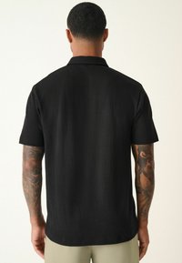 Sort t-shirt med kort ærme og struktureret mønster, lige kant og flad krave, båret med lyse bukser.