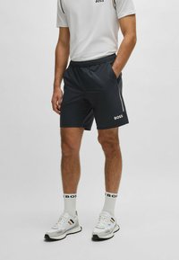 Tamsiai mėlynos sportinės šortai su elastinga juosmena, šoninėmis kišenėmis ir baltu prekių ženklu. Dėvimi su baltu marškinėliu, baltais kojiniais ir sportiniais bateliais.