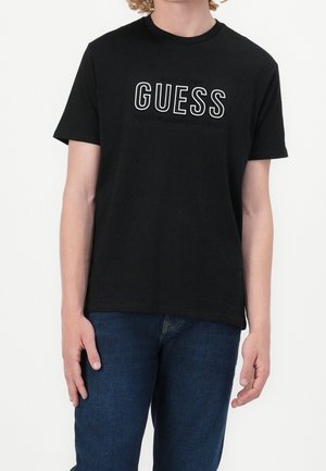 Osoba nosząca czarną koszulkę z krótkim rękawem z logo „GUESS” w białych konturowanych literach, połączoną z ciemnoniebieskimi dżinsami.