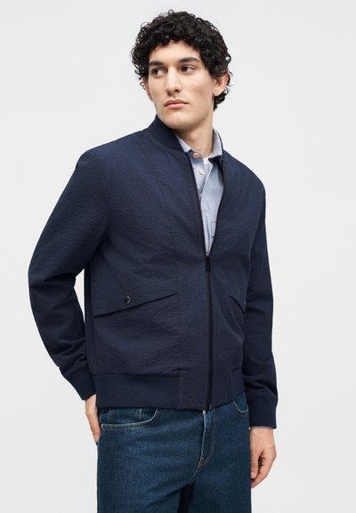 Jeune homme aux cheveux bouclés portant une veste zippée bleu marine par-dessus une chemise bleu clair et un jean bleu, regardant sur le côté.