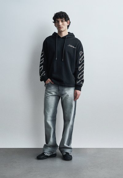 Jeune homme portant un sweat à capuche noir imprimé, un jean gris clair et des baskets noires, debout contre un mur et un sol gris uni.