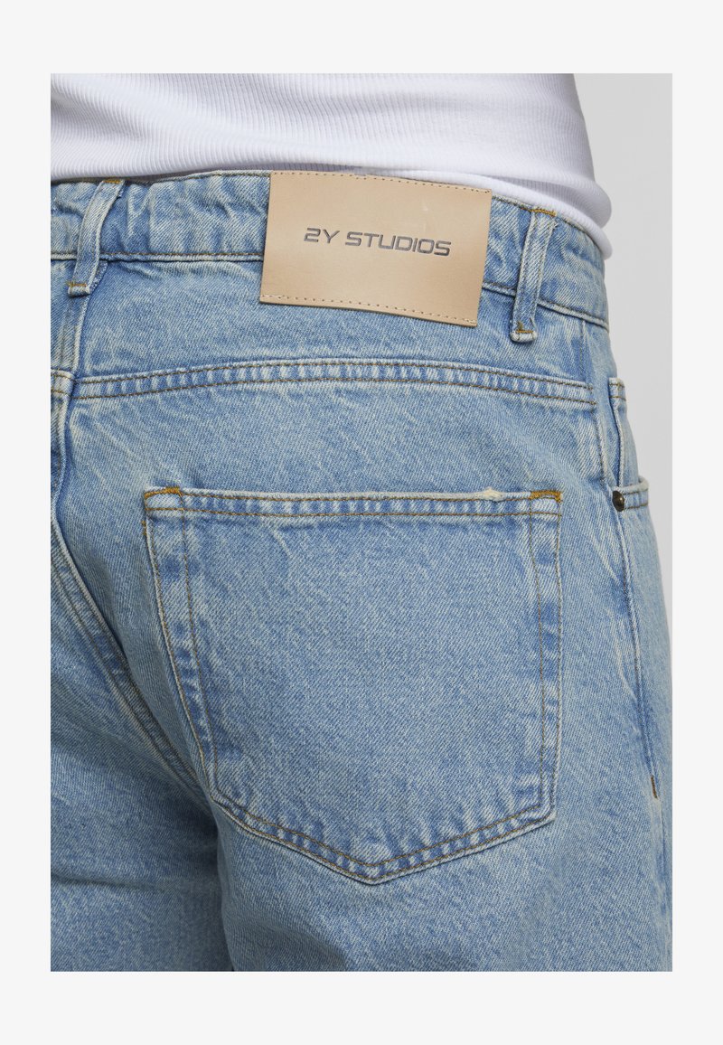 Gros plan sur la poche arrière d'un jean denim bleu clair avec une étiquette beige portant l'inscription "2Y STUDIOS" et une partie d'un t-shirt blanc côtelé rentré à l'intérieur.