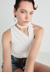 White sleeveless top with a twisted design and a choker detail; worn with distressed denim shorts; model has short, wet-look hair.