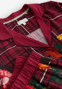 Red and black plaid fabric with floral accents; features a collar, button detail, and gathered texture at the hem. Labeled with brand tag.