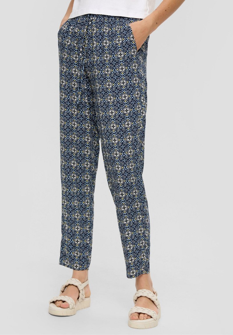 Jambes portant un pantalon bleu à motifs effilés, mains dans les poches et sandales blanches à brides tressées sur fond gris clair.