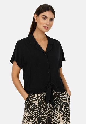 Black short-sleeve button-up blouse with a tied hem detail, made of soft fabric. Paired with patterned light brown and black pants.
