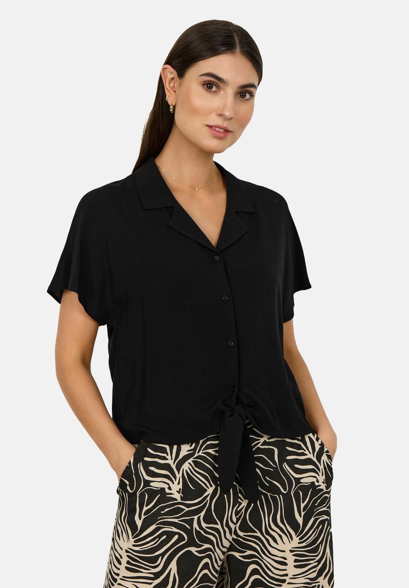 Black short-sleeve button-up blouse with a tied hem detail, made of soft fabric. Paired with patterned light brown and black pants.