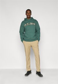 Πράσινο φούτερ με κουκούλα με την επιγραφή "MALBON" σε έντονη, διακοσμητική γραμματοσειρά, συνδυασμένο με μπεζ παντελόνι στενής γραμμής και μαύρα παπούτσια. Απλός, καθημερινός σχεδιασμός.