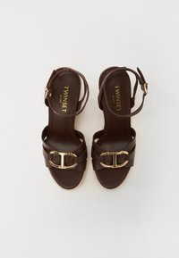 Brown leather sandals with a gold buckle detail, featuring a braided strap design and a natural woven platform sole. Classic ankle strap style.