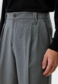 Grijze op maat gemaakte broek van gestructureerde stof met twee voorste knopen en plooien; model gecombineerd met een zwart langmouwtop.