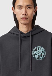 Felpa con cappuccio grigio scuro con coulisse, caratterizzata da un logo grafico circolare blu sul petto. Tessuto morbido, vestibilità casual e tasca frontale.