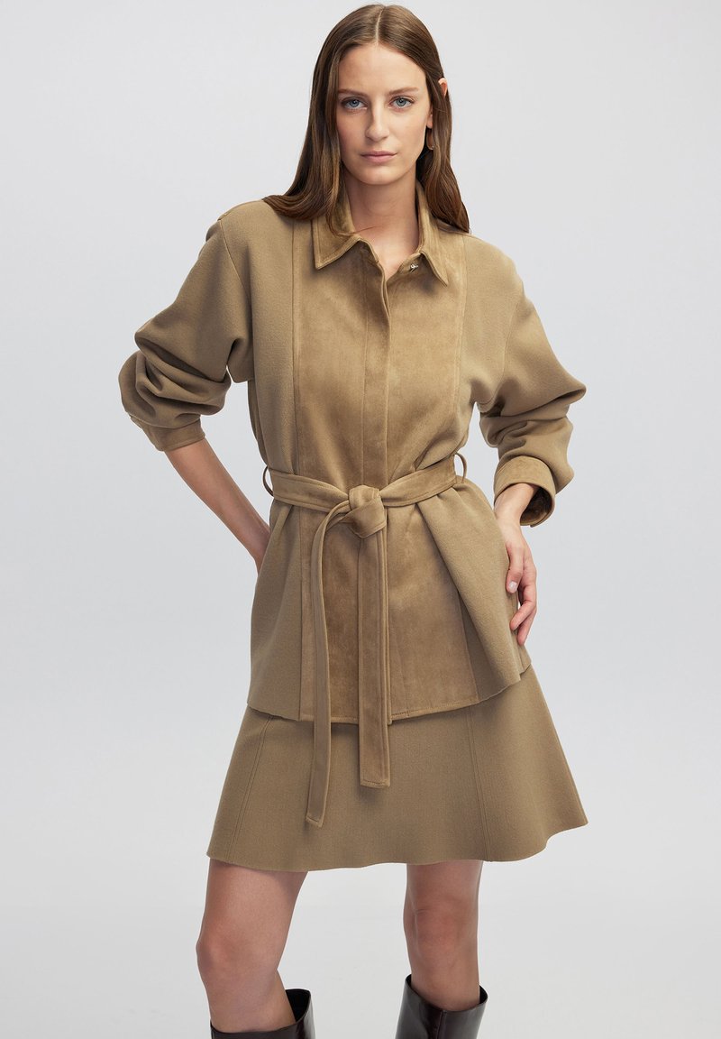 Vestido de ante marrón con cuello abotonado, cintura ceñida con cinturón y falda hasta la rodilla. Mangas largas y abullonadas con textura suave.