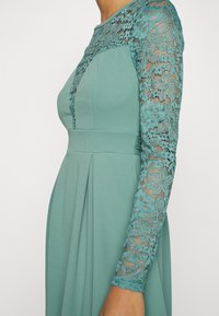 Vestido teal com mangas longas de renda. O corpo é justo, com uma saia plissada. A renda apresenta padrões florais e adiciona textura ao design.