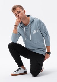 Felpa con cappuccio azzurra chiara con la scritta "WILD" stampata in bianco, abbinata a pantaloni neri slim-fit e sneakers bianche con lacci neri e suole marroni.