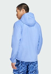 Veste légère bleu clair avec capuche, poignets élastiques et texture lisse. Comprend un accent blanc sur la manche gauche.